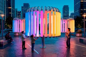 Families interact with the "Pulse of Light" installation in a contemporary town square, gathering around a circular arrangement of vertically suspended glowing tubes.
