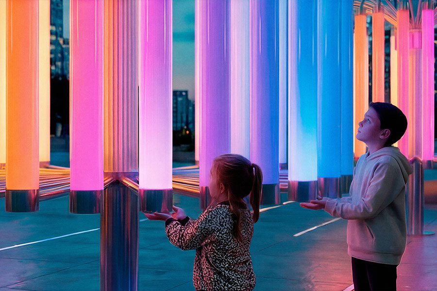 Two children stand beneath the glowing tubes of the “Pulse of Light” installation, holding their hands close to the suspended polycarbonate elements as soft gradients of pink, blue, and warm light shift in response. 