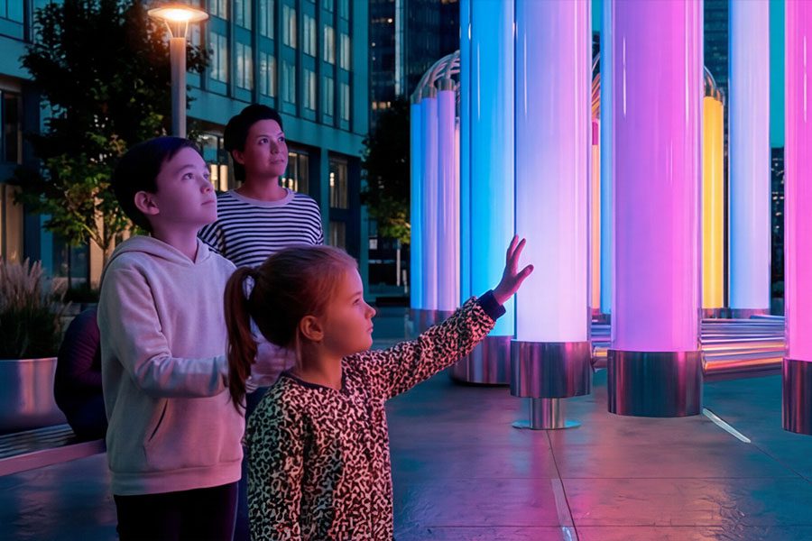 Children interact with the “Pulse of Light” installation in an urban plaza, reaching toward tall illuminated polycarbonate columns that glow with shifting colors. 